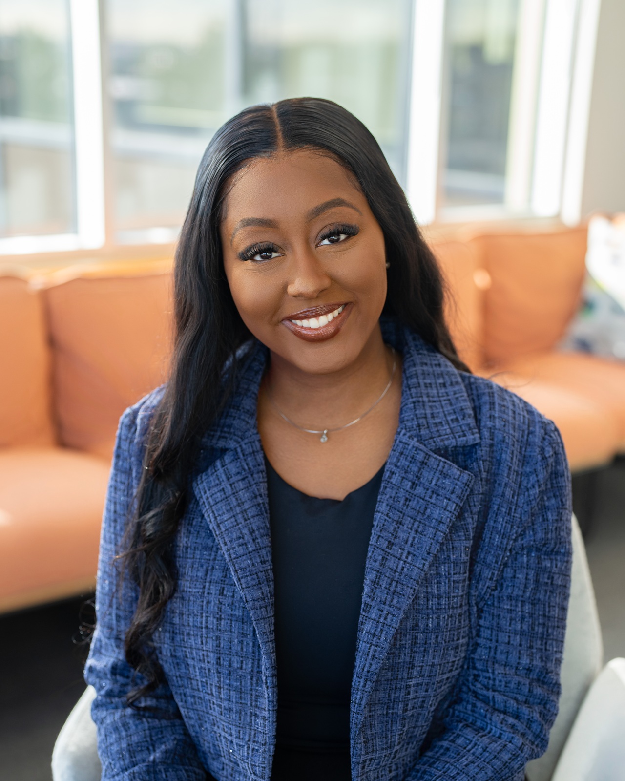 A woman wearing a blue blazer smiling with a window and orange sofa in the background.