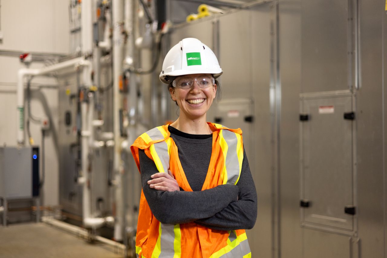 A woman standing in a warehouse and wearing a safety vest, hard hat and goggles.